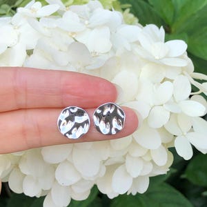 May include: A pair of silver hammered circle stud earrings. The earrings are shown on a finger against a background of white flowers.