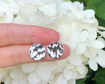 May include: A pair of silver hammered circle stud earrings. The earrings are shown on a finger against a background of white flowers.