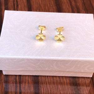 May include: A pair of gold-colored clover stud earrings. The earrings are displayed on a white textured box, set against a dark wood surface. The clover design adds a touch of nature-inspired elegance.
