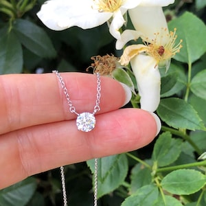 Peut inclure: Un collier délicat en chaîne argentée avec un pendentif rond en pierre précieuse transparente. Le collier est présenté sur un fond de feuilles vertes et de fleurs blanches.