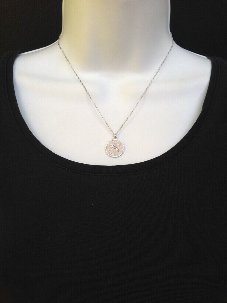May include: A silver necklace with a round pendant featuring a zodiac symbol. The pendant has a textured edge and a delicate chain.