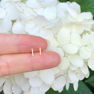 May include: A pair of gold bar stud earrings, shown on a finger against a backdrop of white hydrangea flowers.
