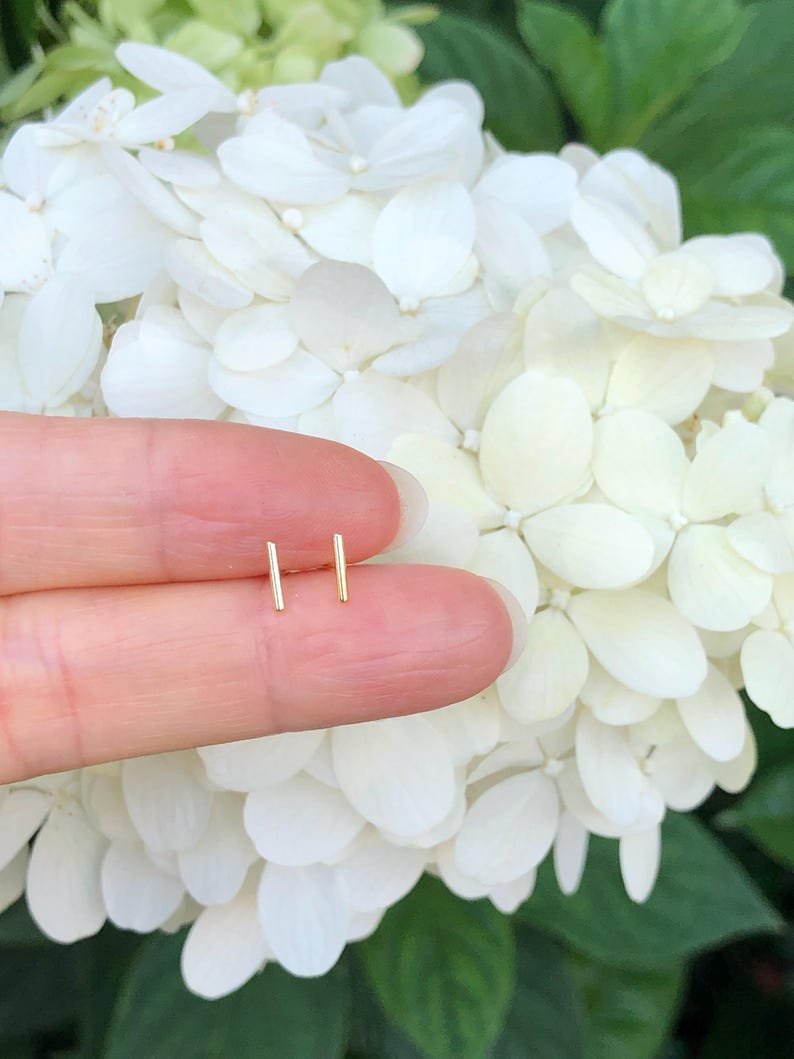 K&ouml;nnte beinhalten: Ein Paar goldene Barren-Ohrstecker, gezeigt auf einem Finger vor einem Hintergrund aus wei&szlig;en Hortensienbl&uuml;ten.
