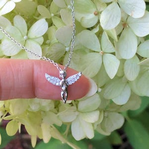 Puede incluir: Un collar de plata con un peque&ntilde;o colgante de &aacute;ngel detallado adornado con piedras transparentes brillantes. El &aacute;ngel tiene las alas extendidas y mira hacia adelante.