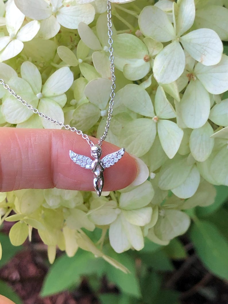 Puede incluir: Un collar de plata con un peque&ntilde;o colgante de &aacute;ngel detallado adornado con piedras transparentes brillantes. El &aacute;ngel tiene las alas extendidas y mira hacia adelante.