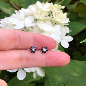 May include: A pair of small, flower-shaped stud earrings. Each earring features a black gemstone center surrounded by white gemstones, set in a silver-colored metal. The earrings are displayed against a backdrop of white flowers and green foliage.