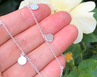 May include: A silver chain necklace with multiple small, round silver charms hanging from it. The necklace is on a hand, and the charms are reflecting light.