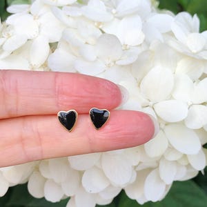 May include: A pair of gold heart-shaped earrings with black enamel. The earrings are small and delicate.