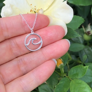 May include: A silver necklace with a delicate wave design pendant. The pendant is a circle with a wave cut out of the center. The necklace is on a hand against a background of green leaves and a white flower.