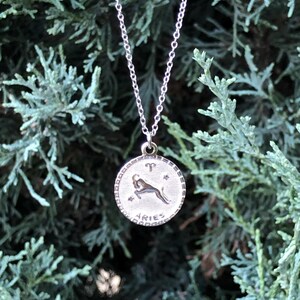 May include: Silver necklace with a round pendant featuring the zodiac symbol for Aries. The pendant has a detailed design of a ram with stars and the word "Aries" inscribed.