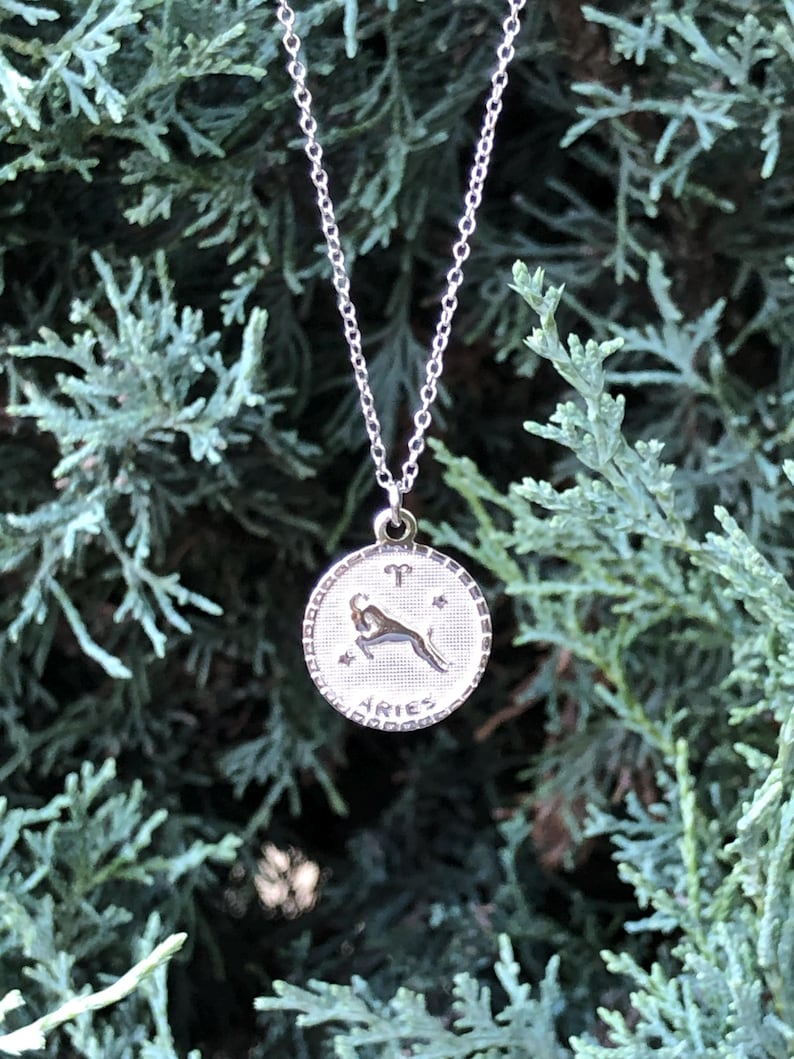 May include: Silver necklace with a round pendant featuring the zodiac symbol for Aries. The pendant has a detailed design of a ram with stars and the word "Aries" inscribed.
