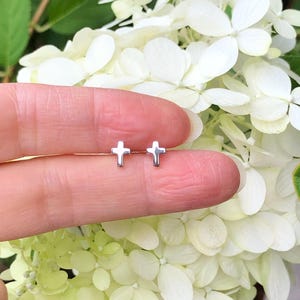 May include: A pair of small silver cross earrings. The earrings are shown on a finger against a background of white flowers.