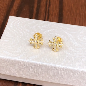 May include: A pair of gold-colored stud earrings. Each earring features a cross design with a central clear stone and smaller stones outlining the cross. The earrings are displayed on a white textured surface.