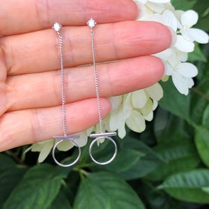 May include: A pair of silver dangle earrings with a small clear stone at the top and a small circle at the bottom. The earrings are hanging from a person's ear.