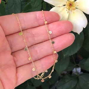 May include: A delicate gold chain necklace with small, round charms hanging from it. The necklace is shown on a hand against a blurred background of green leaves and a white flower.