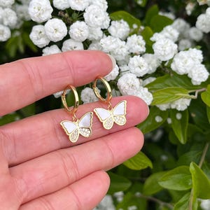 May include: A pair of gold hoop earrings with white butterfly pendants. The butterfly wings have a pearlescent finish and are accented with small, sparkling crystals. The earrings are displayed against a backdrop of white flowers and green foliage.