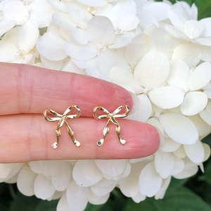 Puede incluir: Un par de pendientes de oro en forma de lazo. Los pendientes son peque&ntilde;os y delicados, y se muestran en un dedo sobre un fondo de flores blancas.
