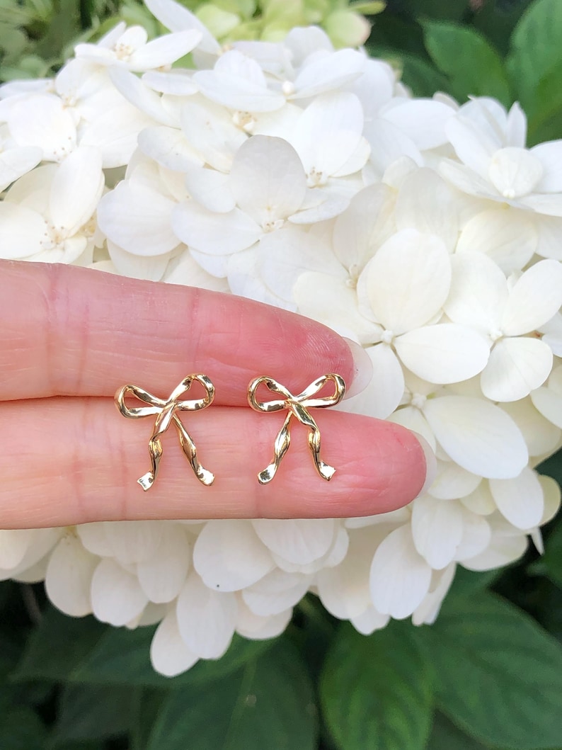 Puede incluir: Un par de pendientes de oro en forma de lazo. Los pendientes son peque&ntilde;os y delicados, y se muestran en un dedo sobre un fondo de flores blancas.