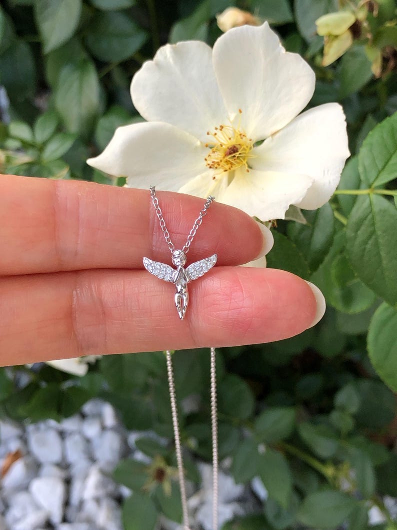 Puede incluir: Un collar de plata con un peque&ntilde;o colgante de &aacute;ngel adornado con piedras de circonita c&uacute;bica transparentes. El &aacute;ngel tiene las alas extendidas y un dise&ntilde;o delicado.