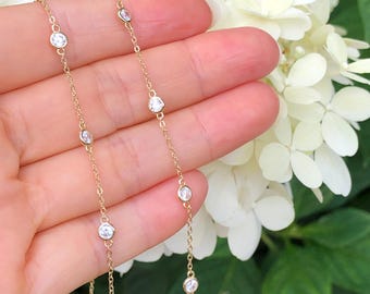 May include: A delicate gold chain necklace with small round clear stones. The necklace is displayed on a hand against a background of white flowers and green leaves.