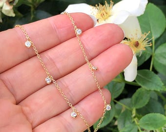 May include: A delicate gold necklace featuring multiple small, clear gemstones. The necklace is displayed against a backdrop of green foliage and white flowers, highlighting its elegant design. The chain is a fine, linked style.