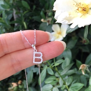 May include: A silver necklace with a letter B pendant. The pendant is made of small, clear stones that form the letter B. The necklace is on a white background.