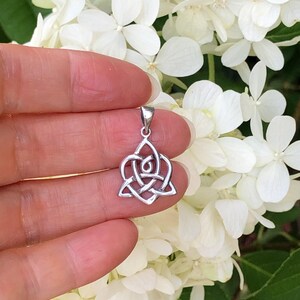 May include: A silver Celtic knot pendant in the shape of a heart. The pendant is small and delicate, and it is held in a hand against a background of white flowers.