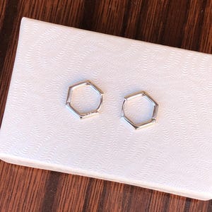 May include: A pair of silver hexagonal hoop earrings. The earrings are on a white background.