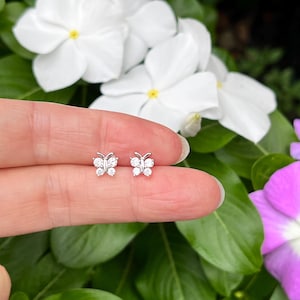 May include: A pair of silver-toned butterfly stud earrings, each featuring four clear, round-cut stones arranged to form the wings, with a small metal body detail in the center, presented as a set of two matching pieces.
