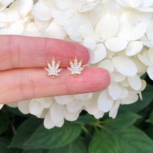 May include: A pair of gold earrings shaped like marijuana leaves, each leaf is encrusted with small clear rhinestones.