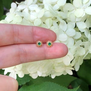 May include: A pair of gold-colored stud earrings. Each earring features a small, round, green gemstone at the center, surrounded by a sunburst design. The earrings are held against a backdrop of white flowers and green foliage.