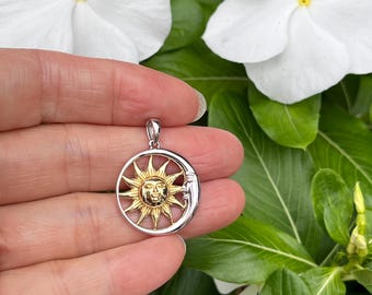Puede incluir: Un colgante circular que presenta un sol dorado con un rostro en el centro, parcialmente rodeado por una luna creciente plateada con un rostro. El colgante incluye una pequeña argolla plateada en la parte superior para una cadena.