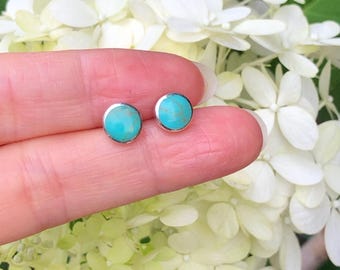 Puede incluir: Un par de pendientes de plata con piedras turquesa. Los pendientes son pequeños y redondos, y las piedras turquesa son de un color azul brillante.