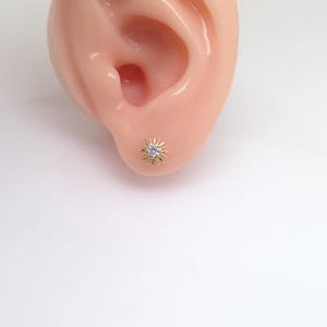 Peut inclure: Boucle d'oreille &agrave; clou dor&eacute;e avec un motif de soleil, orn&eacute;e d'une petite pierre pr&eacute;cieuse transparente au centre. La boucle d'oreille est mise en valeur sur un fond clair, soulignant ses d&eacute;tails et son aspect rayonnant.
