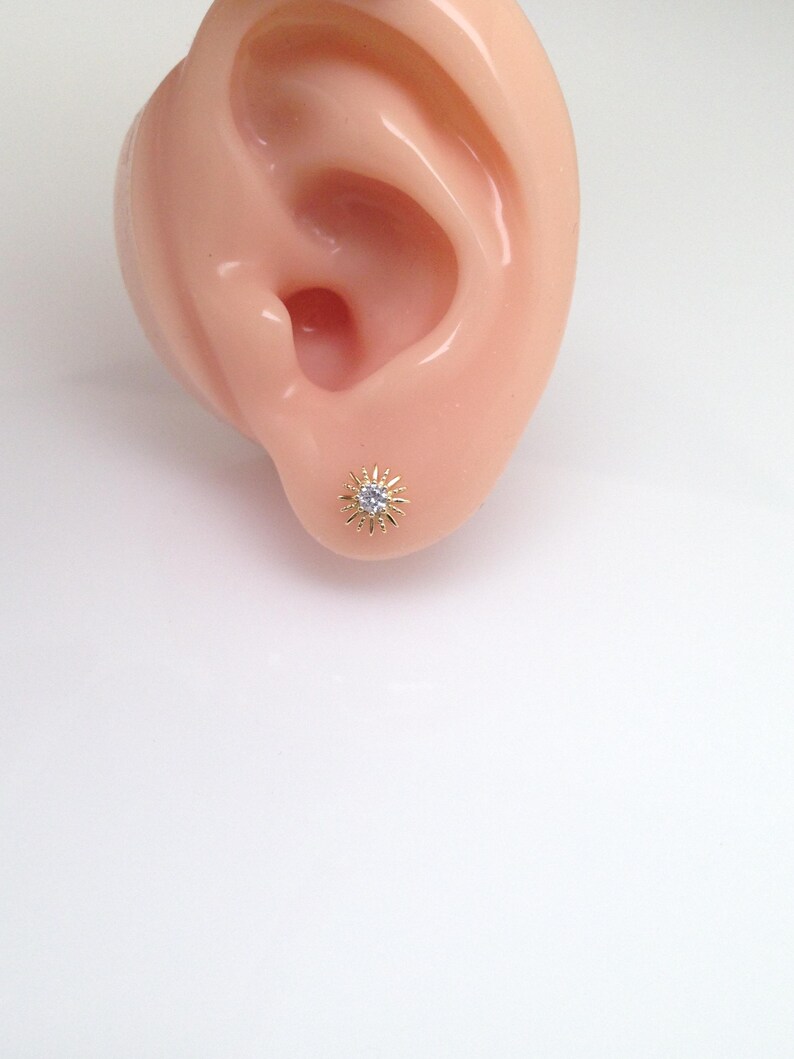 Peut inclure: Boucle d'oreille &agrave; clou dor&eacute;e avec un motif de soleil, orn&eacute;e d'une petite pierre pr&eacute;cieuse transparente au centre. La boucle d'oreille est mise en valeur sur un fond clair, soulignant ses d&eacute;tails et son aspect rayonnant.