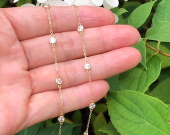 May include: A delicate gold necklace featuring multiple clear, round gemstones. The necklace is displayed against a backdrop of white flowers and green foliage, creating a soft, natural aesthetic. The chain is thin and the gemstones are evenly spaced.