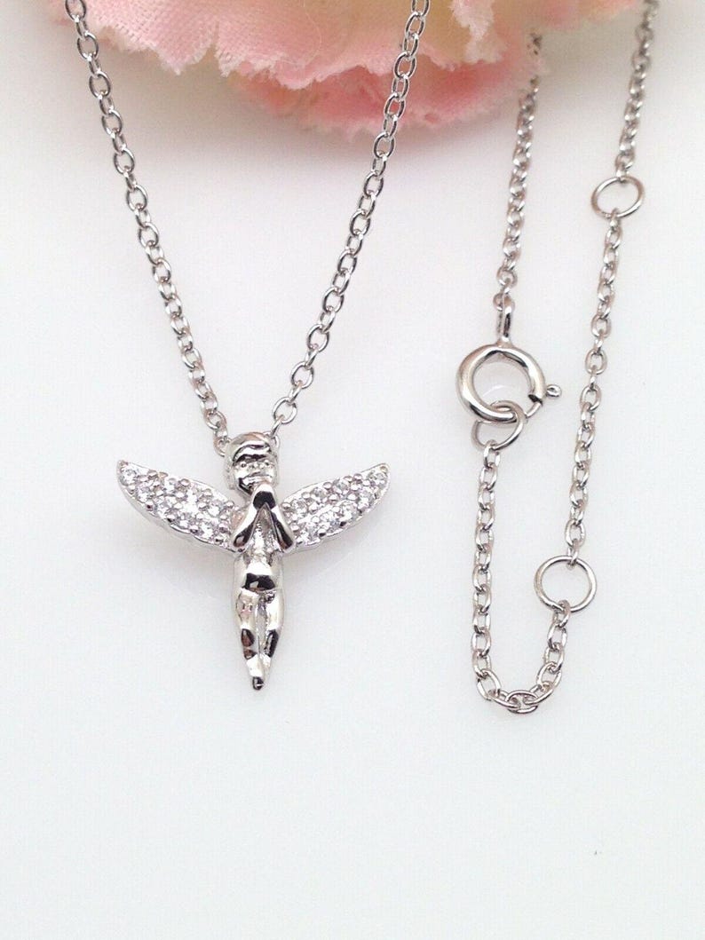 Puede incluir: Un collar de plata con un peque&ntilde;o colgante de &aacute;ngel. El &aacute;ngel tiene las manos juntas en oraci&oacute;n y tiene un peque&ntilde;o halo. El collar est&aacute; sobre un fondo blanco.