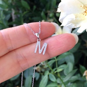 May include: A silver necklace with a pendant in the shape of the letter M. The pendant is encrusted with small, clear stones.