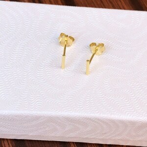 K&ouml;nnte beinhalten: Ein Paar goldene Barren-Ohrstecker in einer wei&szlig;en Schachtel. Die Ohrringe sind schlicht und elegant.