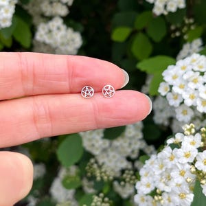 May include: A pair of small, circular silver-toned stud earrings, each featuring a cut-out pentagram design enclosed within a thin metal ring. The earrings have a polished, metallic finish and a simple, symmetrical geometric structure.