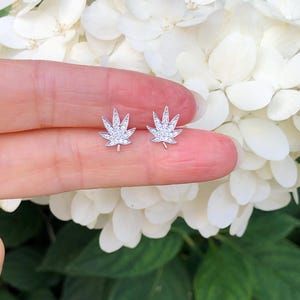 May include: A pair of silver-colored stud earrings shaped like cannabis leaves, each encrusted with small, sparkling stones. The earrings are displayed against a backdrop of white flowers and green foliage.
