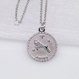 May include: Silver necklace with a round pendant featuring the zodiac sign Aries. The pendant has a textured surface and the word "Aries" is inscribed on the bottom.