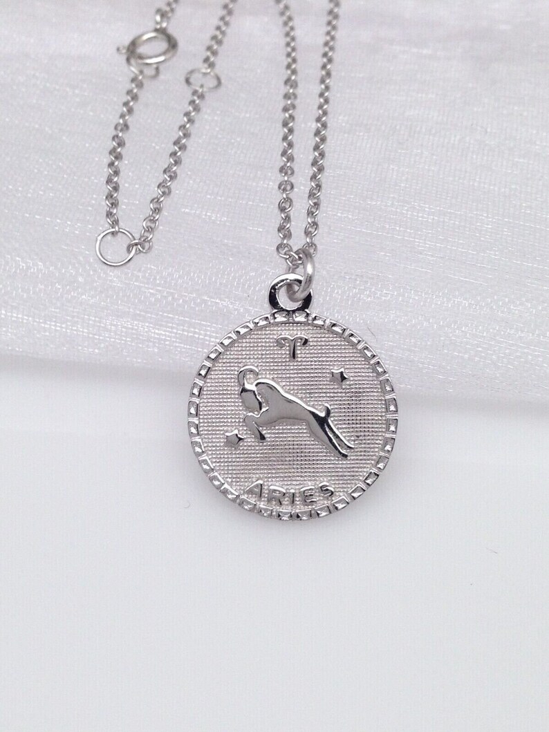 May include: Silver necklace with a round pendant featuring the zodiac sign Aries. The pendant has a textured surface and the word "Aries" is inscribed on the bottom.