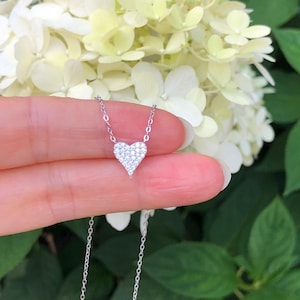 May include: A silver chain necklace with a small, heart-shaped pendant encrusted with tiny clear stones. The necklace is held in a hand against a background of white and green flowers.