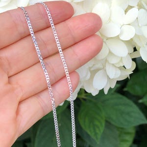 May include: A silver curb chain necklace, shown on a hand against a background of white flowers.