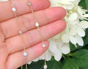 May include: A delicate gold necklace featuring multiple small, round, clear gemstones. The necklace is held against a backdrop of white flowers and green foliage, highlighting the jewelry's sparkle and design. The chain is thin and elegant.