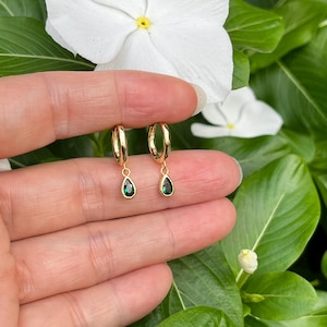 May include: A pair of gold hoop earrings featuring small, dangling, teardrop-shaped charms with dark green stones. The earrings are held against a background of white flowers and green leaves, showcasing the polished metal finish.