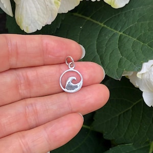 May include: A silver charm in the shape of a wave inside a circle. The charm is being held in a hand.