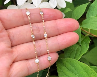 May include: A delicate gold necklace featuring multiple clear, round gemstones. The necklace is displayed against a backdrop of white flowers and green foliage, creating a natural and elegant aesthetic. The gemstones are evenly spaced along the chain.