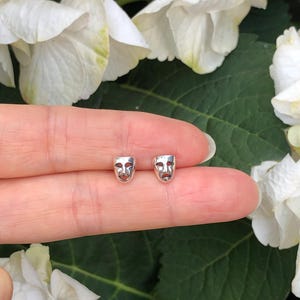May include: A pair of silver earrings featuring a small, detailed design of a theater mask. The earrings are shown on a person's finger against a backdrop of white flowers.